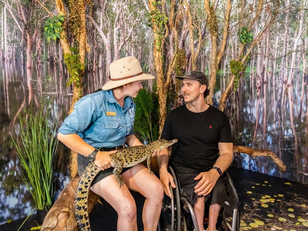 a staff at Crocosaurus Cove holding a baby croc beside a visitor on a wheelchair