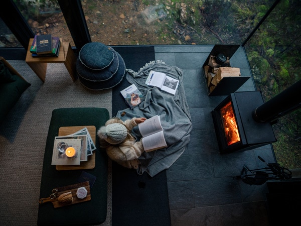 A girl sits by the fire at Pumphouse Point in Tasmania and reads a book. (Image: Emilie Ristevski)