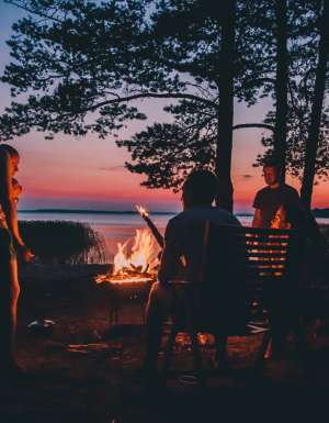 Group of young people near camp fire telling stories near the fire with wood, flames in the nature at night near lake.