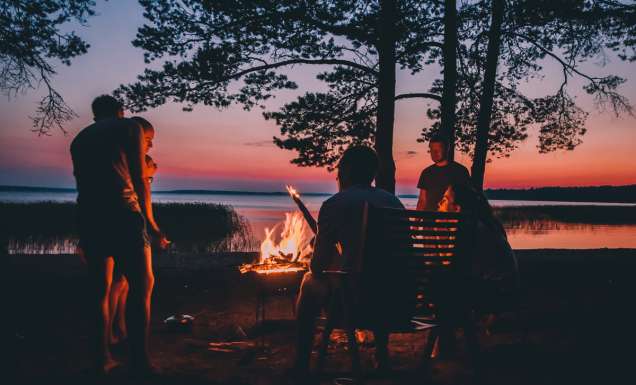 Group of young people near camp fire telling stories near the fire with wood, flames in the nature at night near lake.