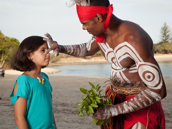 an indigenous member of Yugambeh/Kombumerri people applying white face paint on a kid at Jellurgal Aboriginal Cultural Centre