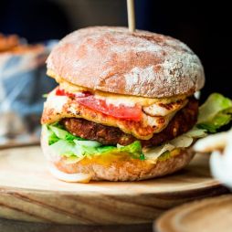 Close up image of a freshly flame grilled vegetarian halloumi cheeseburger on a wooden counter at an outdoor food market. This burger is loaded with a vegetarian burger meat substitute, fresh salad, melted halloumi cheese and spanish onion and tomato. The burger is sandwiched between glazed buns.