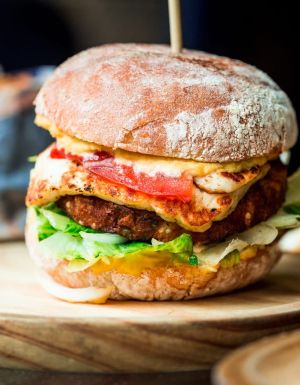 Close up image of a freshly flame grilled vegetarian halloumi cheeseburger on a wooden counter at an outdoor food market. This burger is loaded with a vegetarian burger meat substitute, fresh salad, melted halloumi cheese and spanish onion and tomato. The burger is sandwiched between glazed buns.
