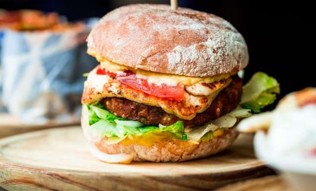 Close up image of a freshly flame grilled vegetarian halloumi cheeseburger on a wooden counter at an outdoor food market. This burger is loaded with a vegetarian burger meat substitute, fresh salad, melted halloumi cheese and spanish onion and tomato. The burger is sandwiched between glazed buns.
