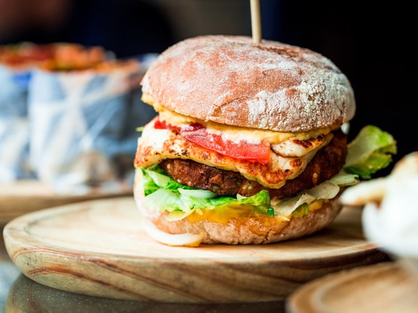 Close up image of a freshly flame grilled vegetarian halloumi cheeseburger on a wooden counter at an outdoor food market. This burger is loaded with a vegetarian burger meat substitute, fresh salad, melted halloumi cheese and spanish onion and tomato. The burger is sandwiched between glazed buns.