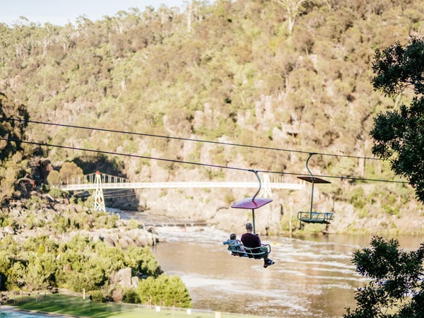 A man and a child sitting and riding a chairlift in the middle of Cataract Gorge reserve and admiring the view overlooking the bridge and mountains.