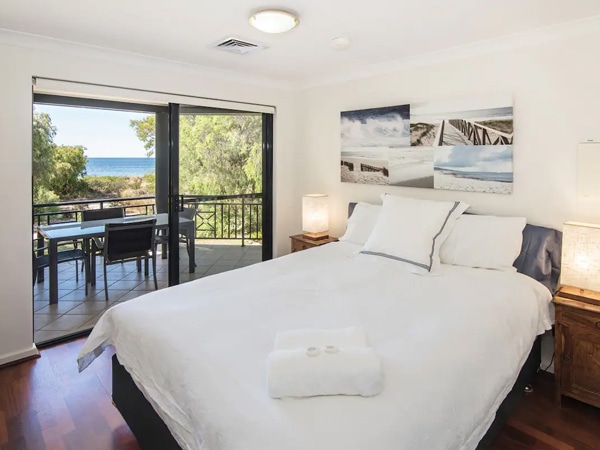 A bed with white sheets and pillows in a bedroom with frames and a balcony with a table and chairs overlooking the ocean.