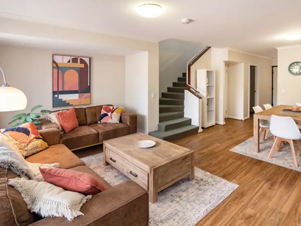 An interior with a couch and pillows, a lamp, a coffee table, a dining table, rugs, and stairs.