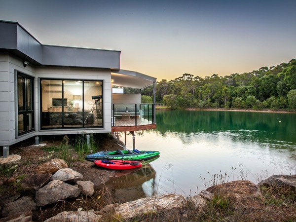 A white house on a lake with kayaks surrounded by trees and rocks.