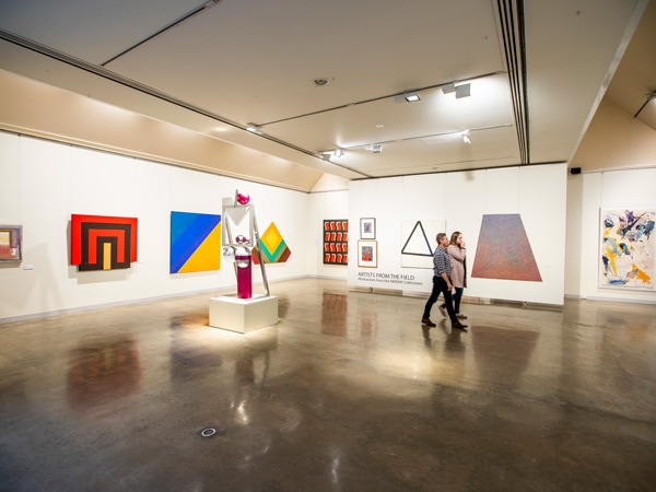 The indoors of a museum with white wall and colorful frames, and two people walking past.