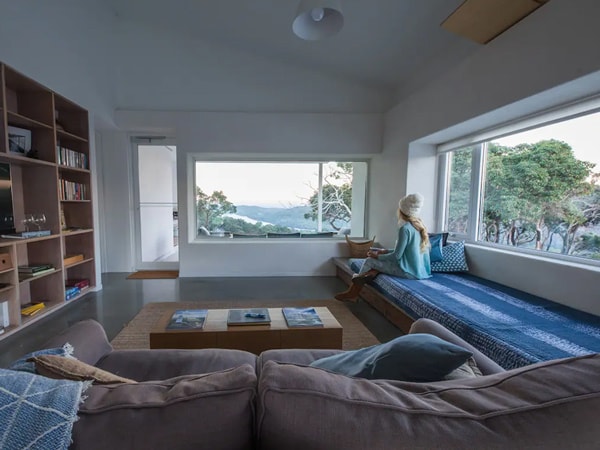 The inside of a house with a girl sitting on a bench looking at the view outside.