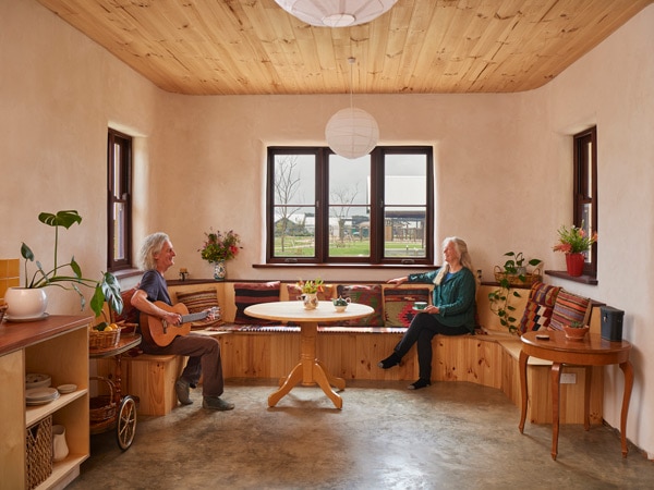 an old man playing guitar with an old lady sitting and looking outside the window