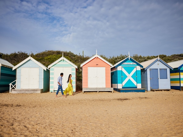 a couple walking along the colourful huts at Brighton Beach, Vic