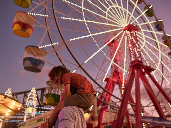 a man giving his child a piggyback ride with the giant glowing Ferris wheel in the background at Luna Park Sydney