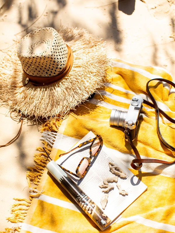 A towel on the beach filled with a hat,,camera and magazine.