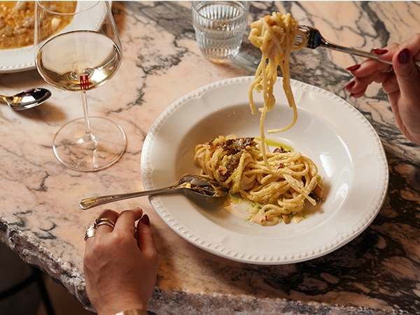 Twirling a plate of pasta over a marble table next to a bowl of risotto and glass of wine.