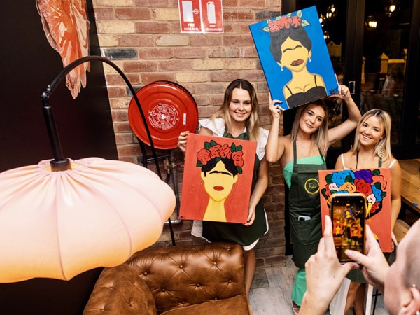 friends posing for a picture while holding their artwork at Frida’s Sip n’ Paint