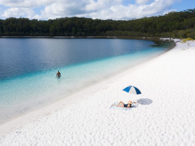 man walks in water while woman relaxes under a beach umbrella in lake mckenzie on k'gari fraser island