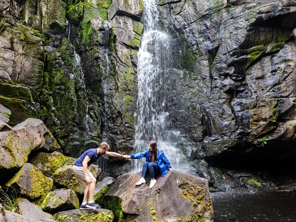 a man reaching out for the hand of the woman sitting on a rock backdropped with the Phantom Falls, Lorne