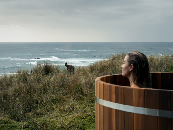 A woman sitting in an outdoor bath looking at a wallaby and the ocean.