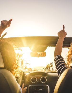 Couple of woman friends traveling and driving having a lot of fun dancing in the car with opened roof and summer vacation sunset ocean in front