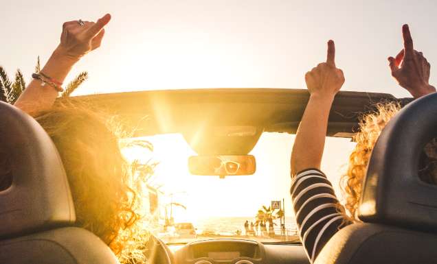 Couple of woman friends traveling and driving having a lot of fun dancing in the car with opened roof and summer vacation sunset ocean in front