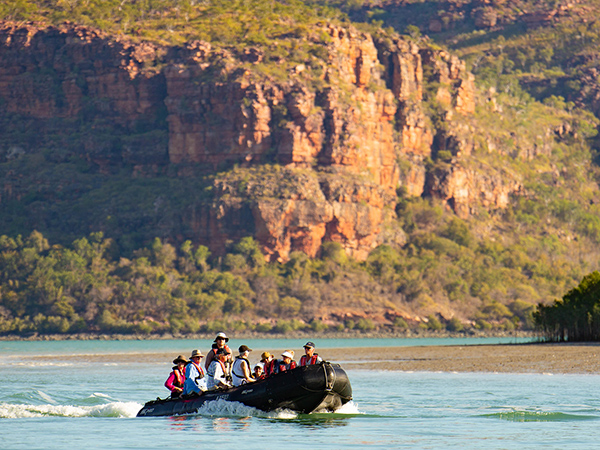 A small group glides through Kimberley’s rugged coastline by boat, passing ancient cliffs.