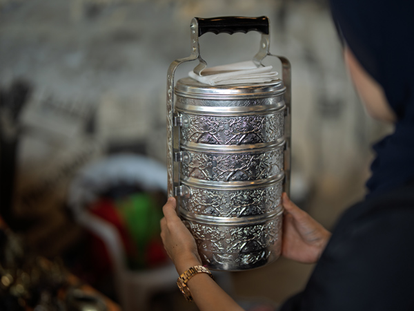 woman wearing a dark outfit holds a traditional embossed silver metal tiffin food carrier with four compartments. The container features intricate floral patterns and a black handle, evoking cultural heritage and craftsmanship