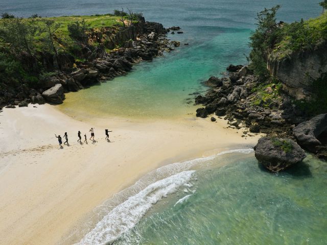 An aerial view of the beach shows tiny figures lined up across the white sand, moving as if in a rhythmic dance.