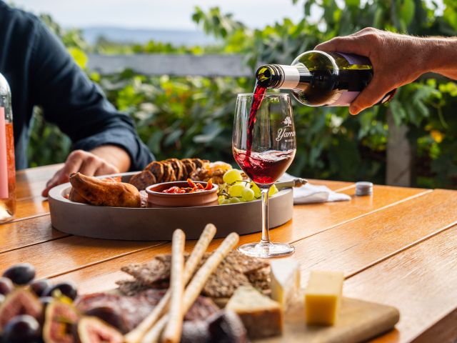 A hand pouring wine into a glass, with a table filled with food.