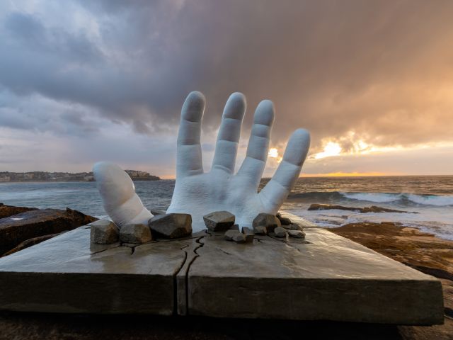 Andre Braun’s giant white hand sculpture Surface Breach rising through a stone slab above the ocean at Sculpture by the Sea, Bondi